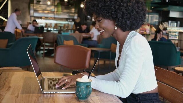 Happy African American woman sitting in cafe drinking coffee and working on laptop and phone, freelance businesswoman video call chatting typing message