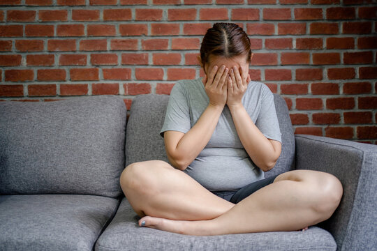 Stressed Fat Woman Sitting On The Sofa In The Living Room, Feeling Disappointed. She Covered Her Face With Her Hands.