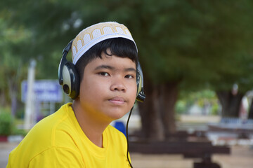 portrait young asian muslim boy in yellow shirt and wears hat, holds headphone and sitting under the tree in the park, soft and selective focus., 