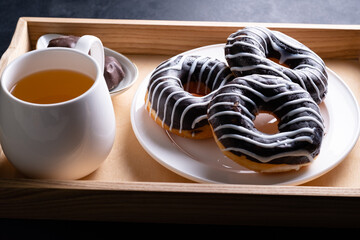 Donut, covered with chocolate glaze, with caramel topping on white plate and cup of tea on a wooden tray.