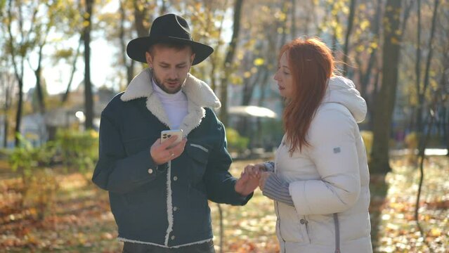 Absorbed Caucasian young man messaging online in smartphone app as mature woman distracting guy. Portrait of concentrated LGBT son scrolling touchscreen as mother talking standing in sunny park
