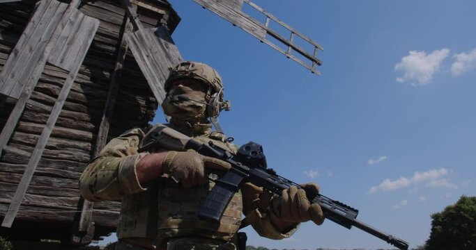 Close up unrecognizable armed soldier in a camouflage uniform standing near a large wooden windmill, World food crisis, war, grain agreement, Ukraine