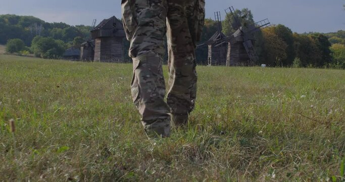 Rear view tracking shot feet of walking unrecognizable armed soldier in a camouflage uniform in the field near a large wooden windmill, World food crisis, war, grain agreement, Ukraine