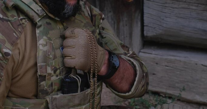 A military man, dressed in a camouflage uniform and tactical gloves, holding a badge and a crucifix on a metal chain in his hand, presses it to his chest, to his heart and praying before the battle.
