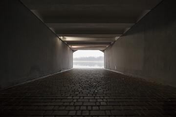 Empty dark underpass tunnel is lit by warm dim lamps, with a view of the foggy river bank in the...