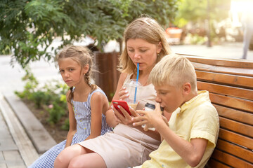 Thoughtful mother, child daughter and teen son having good time together sitting at street with hot drinks on sunny summer day. Happy family sitting on bench using smartphone and have fun together