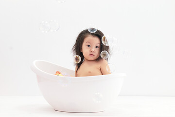 An Asian girl takes a bath with soap bubbles and plays a duck toy. She is sitting in the bath.