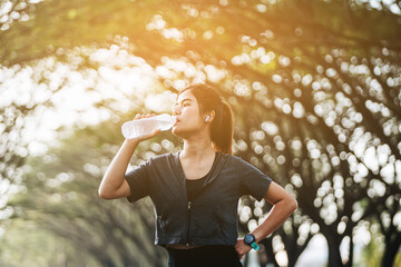 Obraz premium Fitness woman drinking water from sports bottle on afternoon workout after run training jogging outdoors at sunset. Girl wearing wireless headphones silhouette against sun flare.