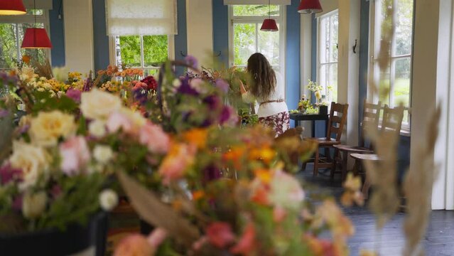 Experienced master florist working near the window in floral studio. Woman white sweater sits at table making wonderful bouquet in spacious light hall. view through flowers on foreground