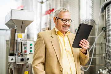 Portrait of happy senior man who owns winery. He is standing beside wine storage tanks and...