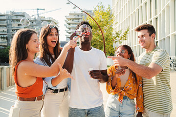 Multiethnic smiling group of teens blowing bubble soup and enjoying outside together next to the campus. Cheerful and young adolescents playing with bubble wand on sunny day in the Street. Lifestyle