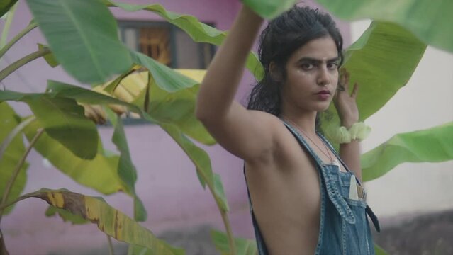 Bold and beautiful south Asian woman looking through banana leaves wearing denim dungarees with revealing upper body parts and bohemian look 