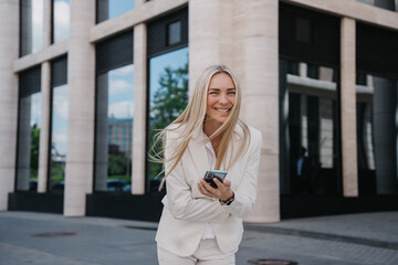 Fototapeta premium Excited young blonde businesswoman in white suit holds phone laughing standing outside against building makes call laughing toothy smiles. Cheerful caucasian female manager received great news.