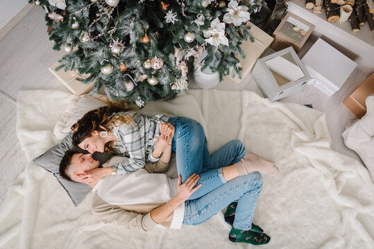Man hugs woman lying down on blanket on floor near Christmas tree in decorated bedroom interior. Couple enjoying winter time together on holidays. Happy New Year. Xmas at home. Top view.