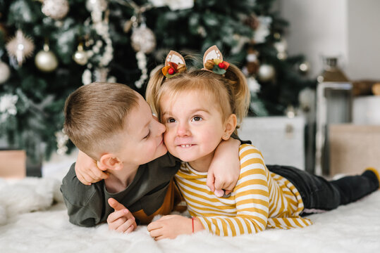 Happy children hugging at a home. Brother kiss sister. Childs playing near Christmas tree. Happy New Year and Merry Christmas. Сoncept of a family holiday. Decorated interior room of a house. Closeup.