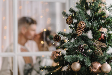 Christmas tree with garlands. Man kissing woman in decorated interior room. Couple enjoying winter time together on holidays. Happy New Year and Merry Christmas. Xmas at home. View through the window.
