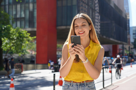 Brazilian girl using mobile phone app for chat on sunny day in Paulista Avenue, Sao Paulo, Brazil