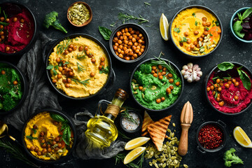 Colorful hummus bowls - green, yellow and beetroot hummus on dark background with lemon, olive oil, and spices. On a concrete black background.