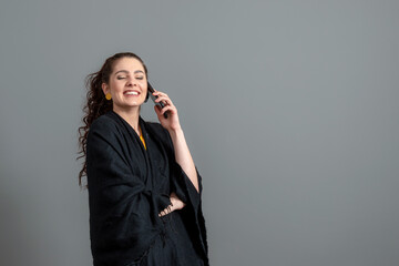 curly-haired emotional young woman in a black cloak talk on mobile phone, isolated on gray