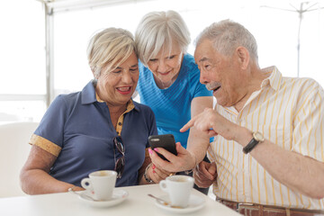 gruppo di tre anziani  sorridenti guarda il cellulare seduti comodamente in un tavolino