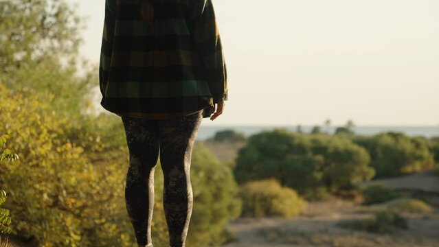 A girl in leggings and a plaid shirt on a high slope with pine trees admires the sunset and the sea.