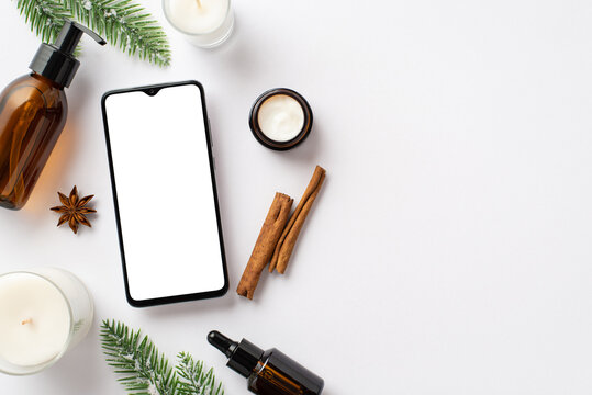 Winter skincare concept. Top view photo of smartphone amber pump bottle cream jar glass dropper bottle fir branches in frost candles cinnamon sticks anise on isolated white background with blank space