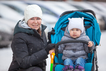Mom in a warm winter jacket and hat walks with a two-year-old child in a stroller
