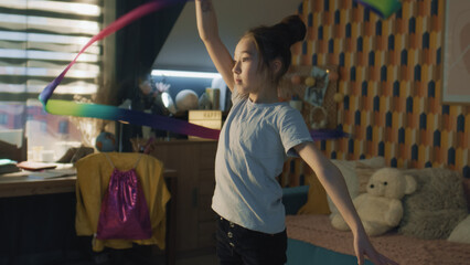 Obraz na płótnie Canvas Young asian schoolgirl spins gymnastic ribbon around herself, and trains new visual moves. Rhythmic gymnastics. Sports hobby. Upgrading physical health on quarantine. Slow motion.