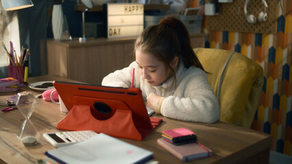 Young asian girl sits at the table at cozy room and talks with teacher. Schoolgirl listens online lesson, studies on tablet,writes down and discuss new material. Distance learning concept.