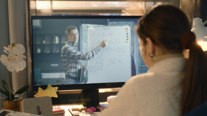 Young asian girl sits at the table at cozy room and learns new material. Schoolgirl listens online lesson of math and studies on pc. Mature teacher talks with student and shows equation on board
