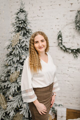 A beautiful young girl is standing near Christmas tree in living room. Christmas tree and home decorated interior. Happy New Year. Fashion woman relaxing in the warm and cozy evening at home. Closeup.