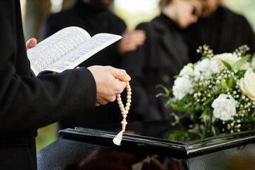 Close up of person in black praying at outdoor funeral and holding Bible with rosary, copy space