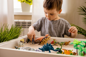 Cute baby boy playing sensory box kinetic sand table with carnivorous and herbivorous dinosaurs - obrazy, fototapety, plakaty