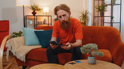 Bearded man using credit bank card and smartphone while transferring money, purchases online shopping, order food delivery at home apartment indoors. Young redhead guy in living room sitting on couch