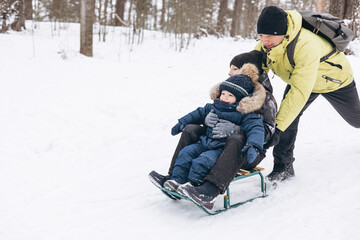 Father with backpack and little sons walking together in winter snowy forest. Happy man and joyful boys sledding and having fun together. Wintertime activity outdoors. Concept of local travel