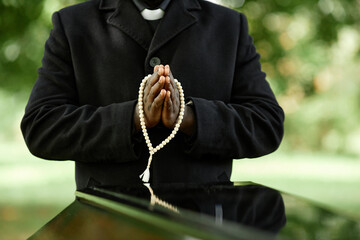 Close up of African American priest wearing black at outdoor funeral ceremony with focus on hands in prayer - obrazy, fototapety, plakaty