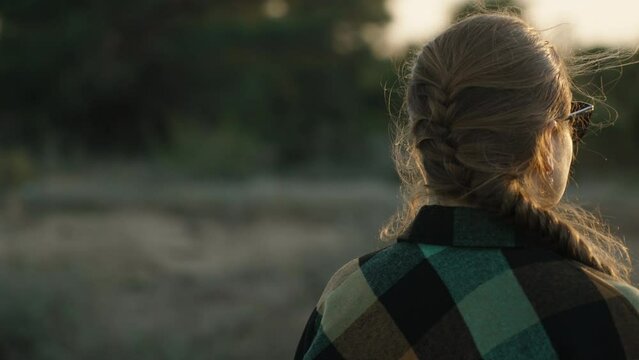 A girl in glasses and a plaid shirt in the forest looks at the sunset, her hair is braided and fluttering in the wind. The camera moves around her.