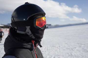 Girl or boy in ski helmet, sunscreen mask and balaclava close up stands against the backdrop of snow-covered mountains and a cloudy sky. Winter. Sport and travel content  