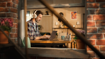 Handsome Caucasian Man Working on Laptop Computer, Sitting Behind His Desk at Home Office. Freelance Entrepreneur Listening to Music, Doing Remote Telework. View From Outdoors into Window.