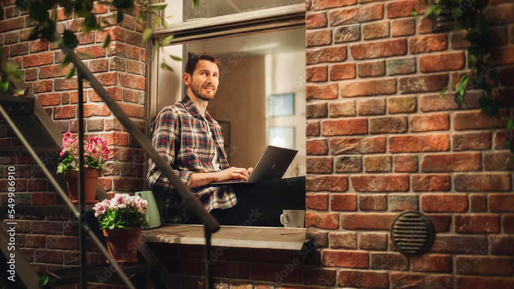 Poster handsome creative male works on a laptop computer and sitting on a windowsill of his cozy apartment.