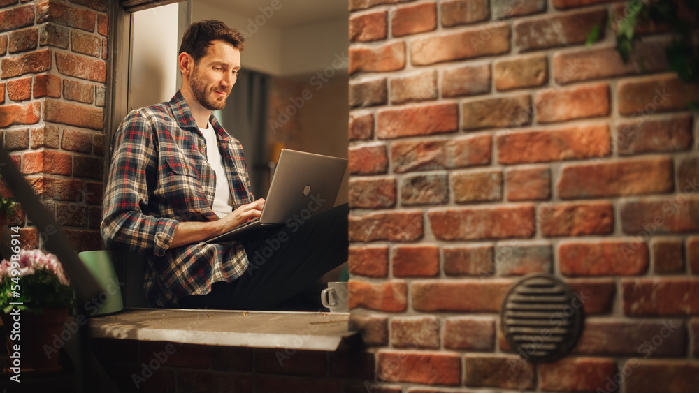 Poster handsome creative male works on a laptop computer while sitting on a windowsill of his cozy apartmen