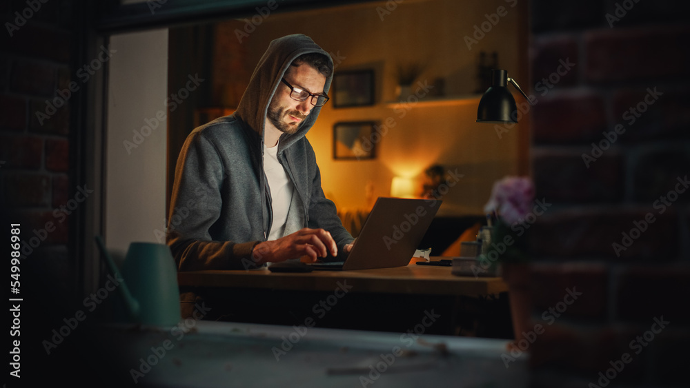 Poster Professional Male Programmer Writing Code for New Project and Working Remotely on Laptop From Home Office. Freelance Software Engineer Designs App From his Apartment. View From Outdoors into Window.