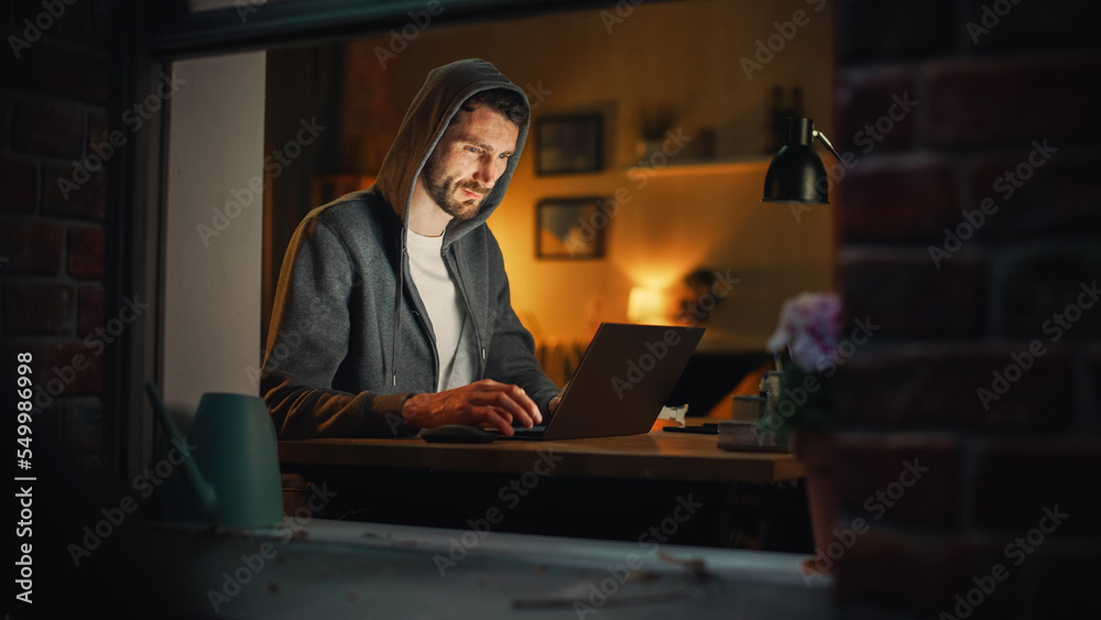 Leinwandbilder Professional Male Programmer Writing Code for Complex Project and Working Remotely on Laptop From Home Office. Freelance Software Engineer Designs App in Apartment. View From Outdoors into Window.