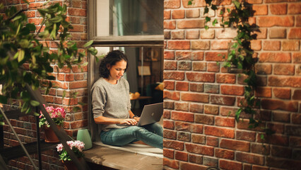 Creative Female Writer Works on a Laptop Computer and Sitting on a Windowsill of her Cozy...