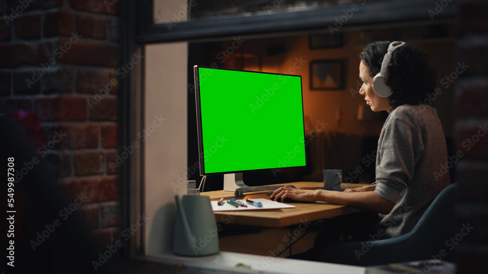 Poster Female Designer Using Desktop Computer with Green Screen Chroma Key Monitor. Remote Access Manager Listening to Music or Podcast in Headphones, Freelancer Working at Night. Into the Window Shot.