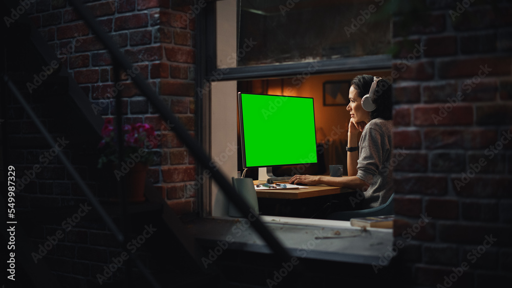 Poster Female Designer Using Desktop Computer with Green Screen Chroma Key Monitor. Remotly Working Manager Listening to Music or Podcast in Headphones, Freelancer Working at Night. Inside the Window Shot.