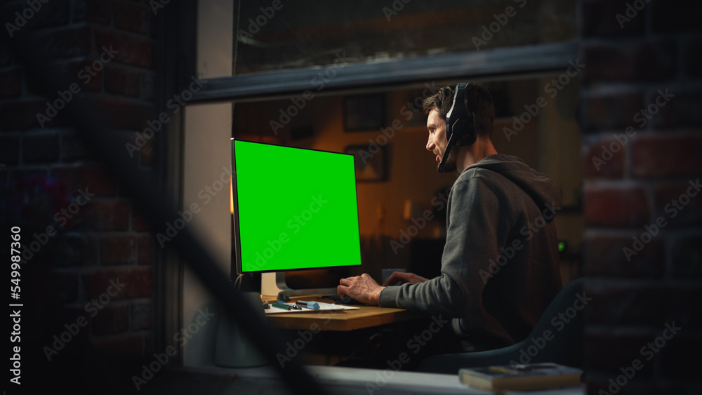 Poster Male Designer Using Desktop Computer with Green Screen Chroma Key Monitor. Remotly Working Manager Listening to Music in Headphones, Freelancer Working at Night. Inside the Window Shot.