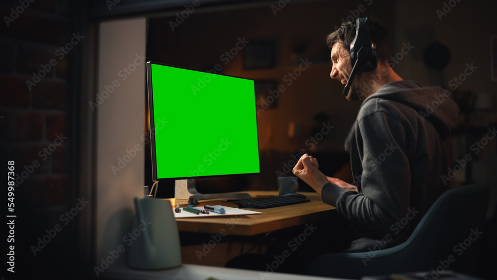 Poster Male Designer Using Desktop Computer with Green Screen Chroma Key Monitor. Remotly Working Manager Listening to Music in Headphones, Freelancer Working Late at Night. Inside the Window Shot.