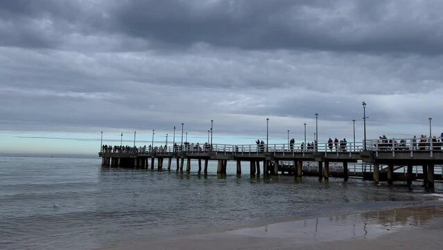 Long wooden pier at the Baltic coast in Sopot, Poland with white railing and street lamps on a cold, cloudy day. Green water and dark sky. High quality 4k footage