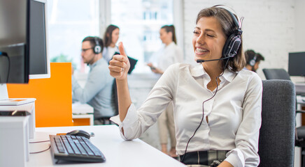 Smiling woman call center operator in headset showing thumbs up gesture to customer in video call.